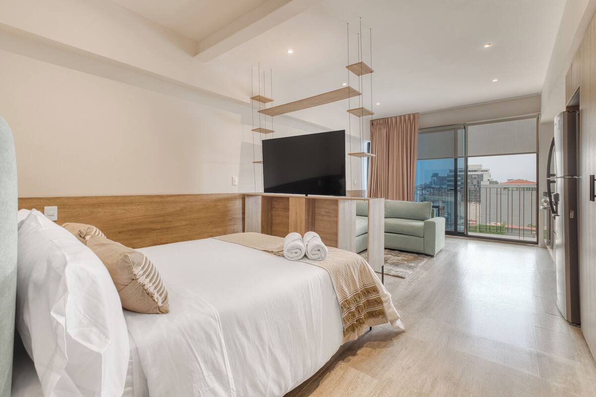 A modern hotel room with a large bed, a flat-screen TV, a balcony with a city view, and a kitchenette.