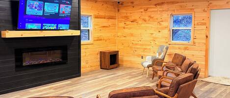 A cozy room with a TV mounted on a black wall, a fireplace, wooden walls, and a seating area with brown chairs.