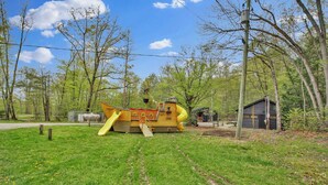 Une aire de jeux avec un toboggan et une structure d’escalade, entourée d’arbres et d’un petit bâtiment.