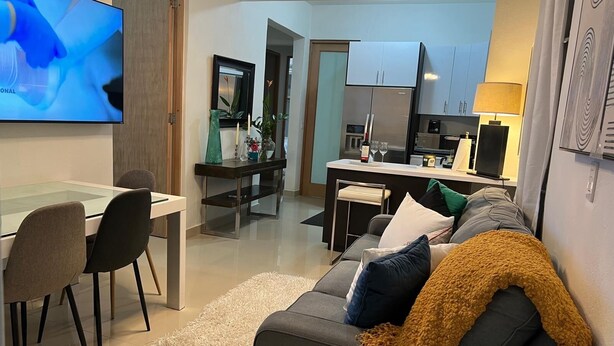 Ein modernes Wohnzimmer mit einem grauen Sofa, einem Couchtisch und einem Fernseher. Im Hintergrund ist eine Küche erkennbar, mit Kühlschrank, Herd und Schränken.