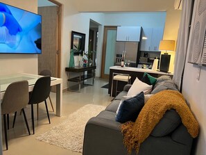 Una sala de estar moderna con un sofá gris, una mesa de centro y un televisor. Se ve una cocina al fondo con un refrigerador, estufa y armarios.