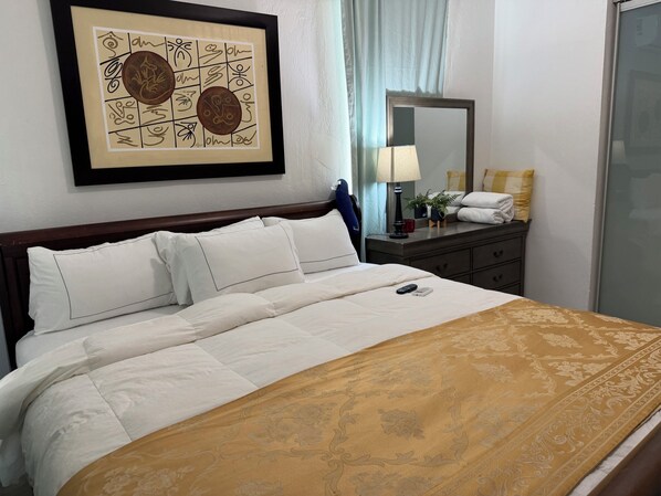 Una cama bien hecha con ropa blanca y un plaid estampado, una mesita de noche con una lámpara, un espejo y una mesita de noche con una planta y una toalla.