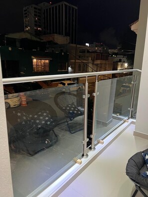 Un balcón con barandilla de vidrio, vistas a coches aparcados y edificios al fondo.