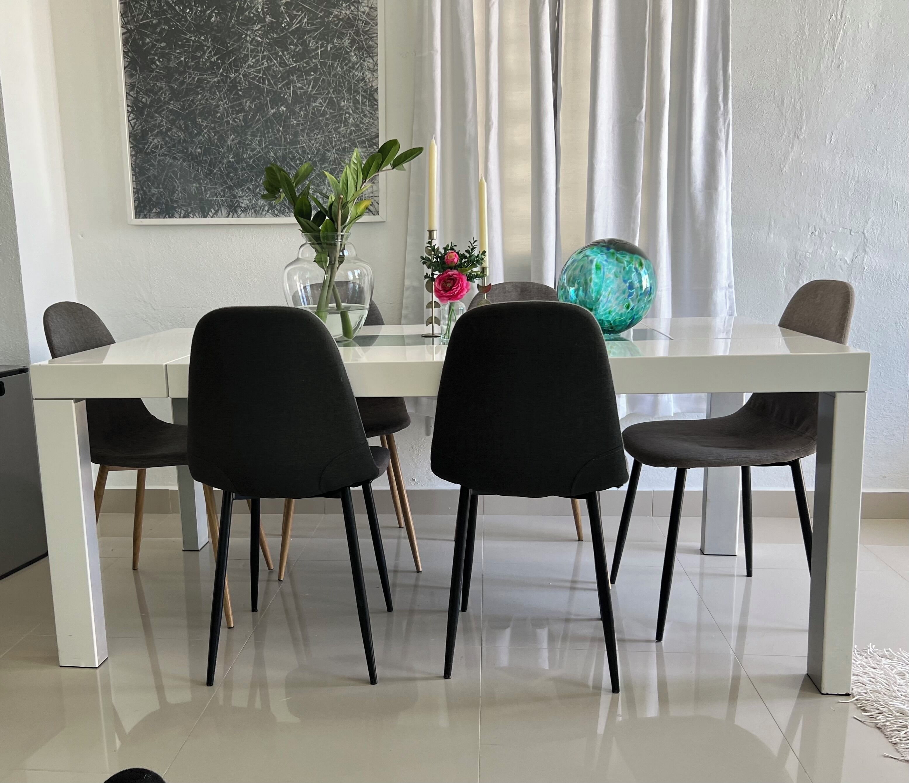 Un espace repas moderne avec une table blanche, des chaises noires, un vase de fleurs et une sphère de verre décorative.