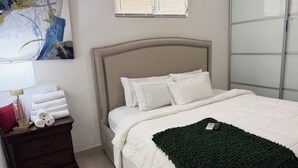 Ein ordentlich bezogenes Bett mit weißer Bettwäsche und Kissen, ein dunkles Holz-Nachttische mit Lampe und Handtüchern sowie eine grüne, gestrickte Decke.