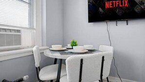 A room with a dining table, white chairs, a small plant, and a wall-mounted TV displaying the Netflix logo. - Bright&inviting~1BR suite~In Uptown (Charlotte)