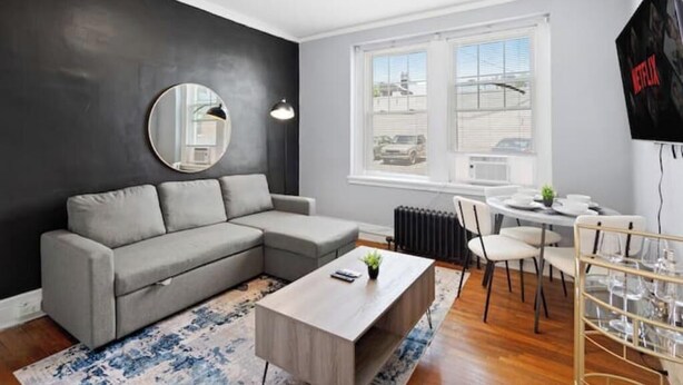 A modern living room with a grey sectional sofa, a round mirror, a wooden coffee table, and a dining area with a white table and chairs. - Bright&inviting~1BR suite~In Uptown (Charlotte)