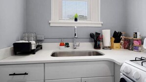 A kitchen with a white countertop, stainless steel sink, and a stove. There is a window above the sink with blinds, and a small plant on the windowsill. - Bright&inviting~1BR suite~In Uptown (Charlotte)