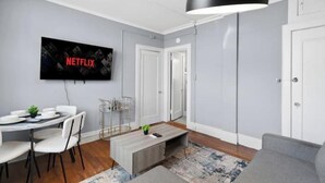 A modern living room with a flat-screen TV mounted on the wall, a grey sofa, a coffee table, and a dining area with a white table and chairs. - Bright&inviting~1BR suite~In Uptown (Charlotte)