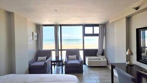 Une chambre d’hôtel avec un lit, deux fauteuils, un bureau et une vue sur la plage.