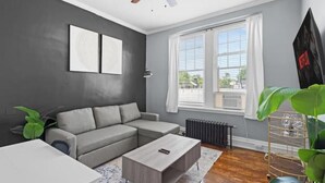 A modern living room with a grey sofa, a wooden coffee table, and a large window with blinds. - Cozy&Central Suite~1BR~In Uptown (Charlotte)