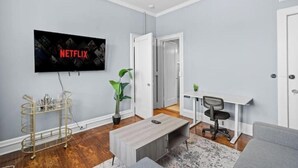 A living room with a flat-screen TV mounted on the wall, a plant, a desk with a chair, and a couch. - Cozy&Central Suite~1BR~In Uptown (Charlotte)