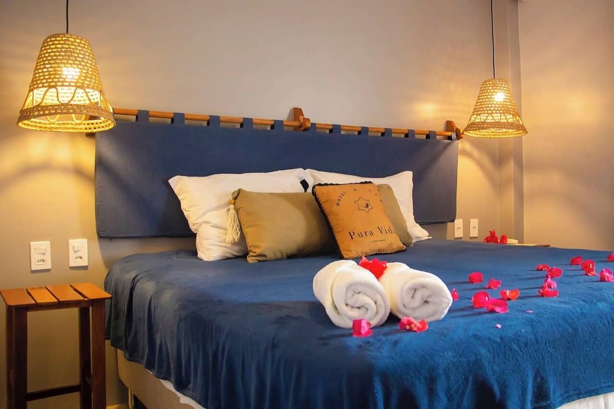 Cama bem arrumada com colcha azul, duas almofadas brancas, uma almofada marrom com o texto 'Pura Vida' e duas toalhas enroladas com flores vermelhas.