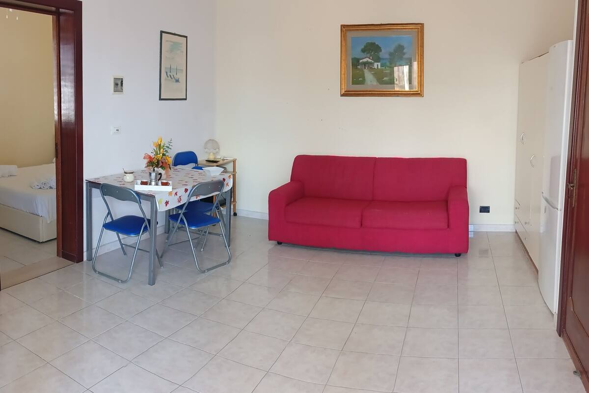 Un salon avec un canapé rouge, une table à manger avec des chaises, une petite table avec une lampe et un tableau accroché au mur.