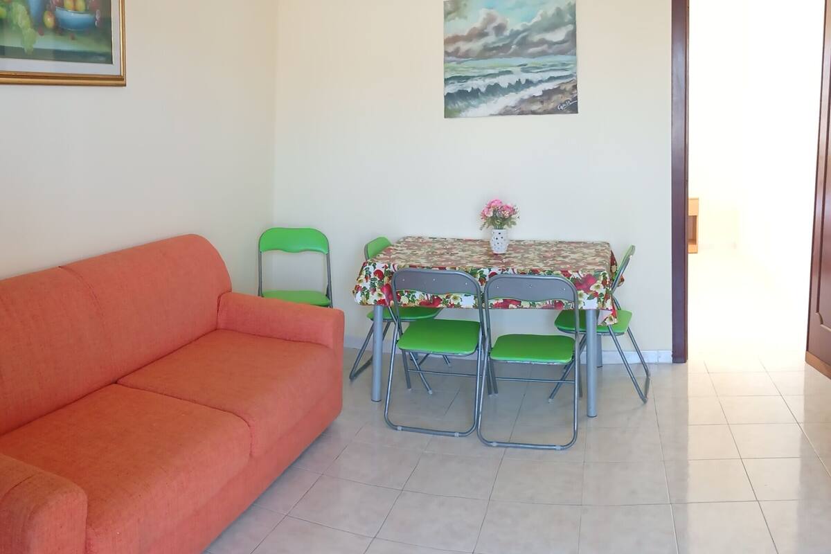 Un salon avec un canapé, une table à manger et des chaises, un tableau représentant un paysage, et une porte donnant sur l’extérieur.