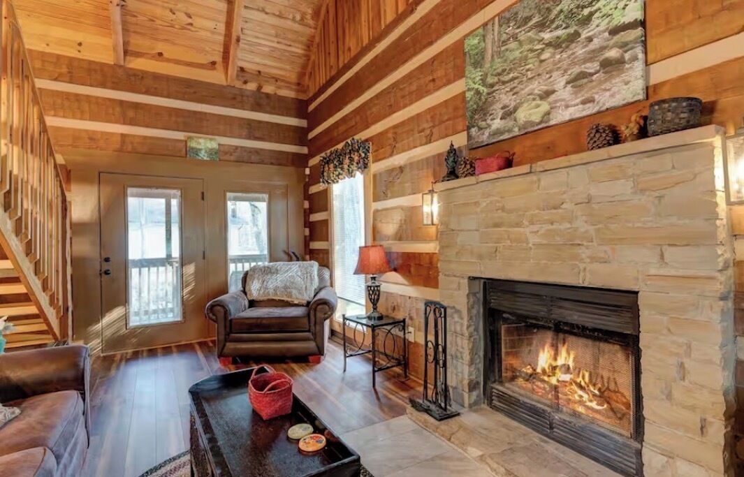 A log cabin interior with a stone fireplace, wooden floors, and a staircase.