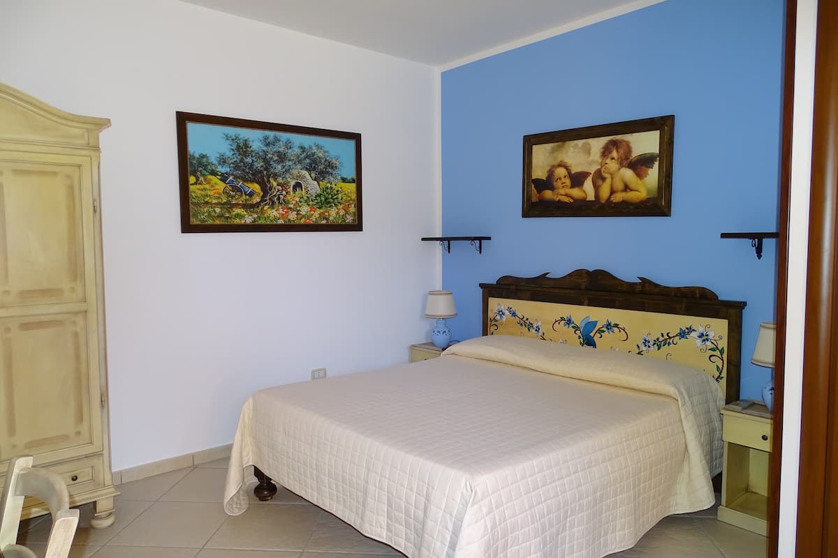 A bedroom with a bed, a nightstand, a lamp, a wardrobe, and two paintings on the wall.