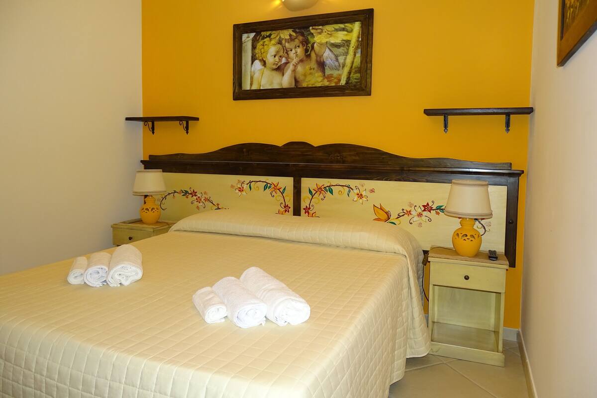 A hotel room with a bed, two bedside tables, a nightstand, a wall-mounted lamp, and a painting above the bed.