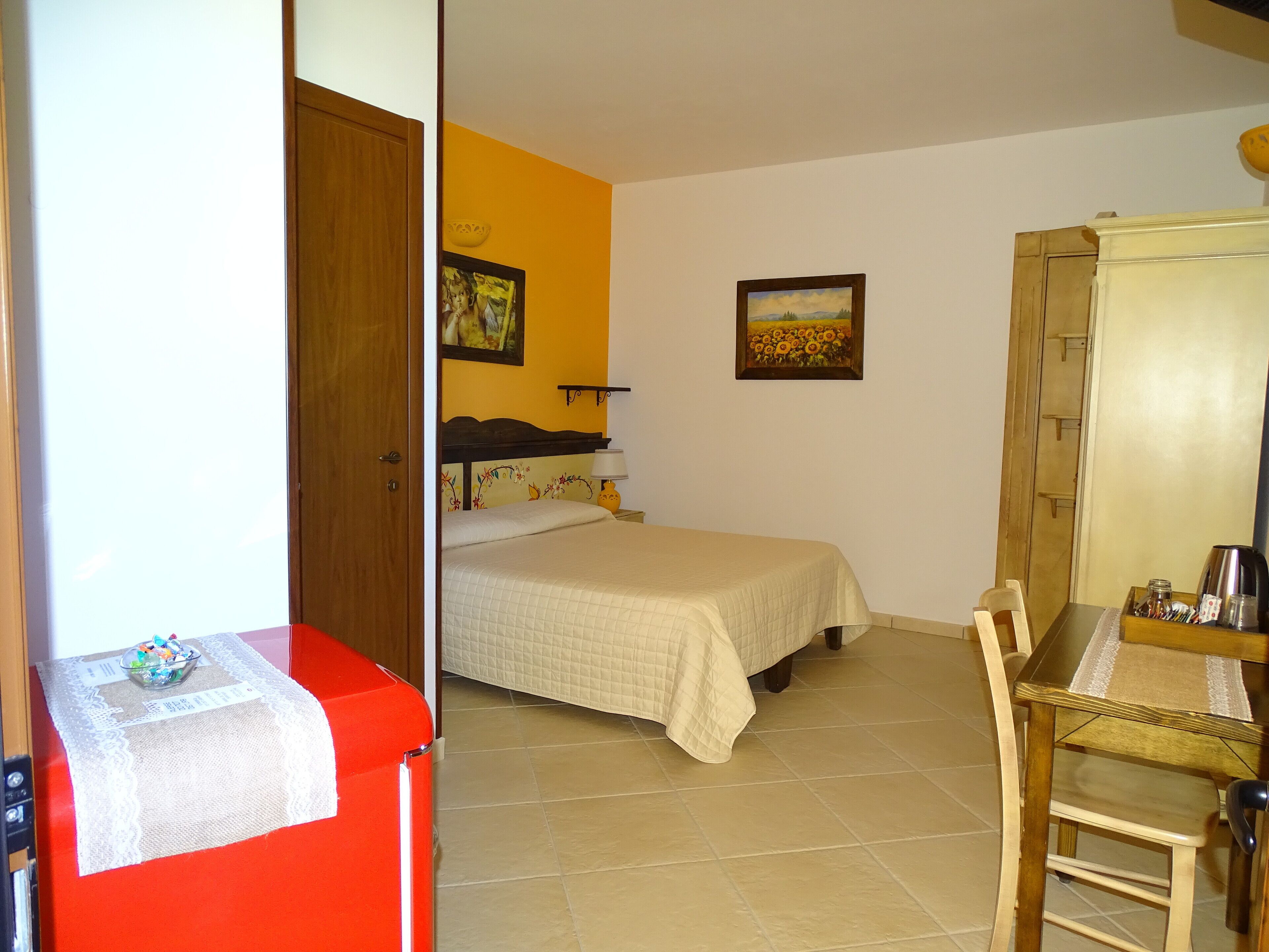 A small, well-lit room with a bed, a wooden wardrobe, a table with a chair, and a red appliance.