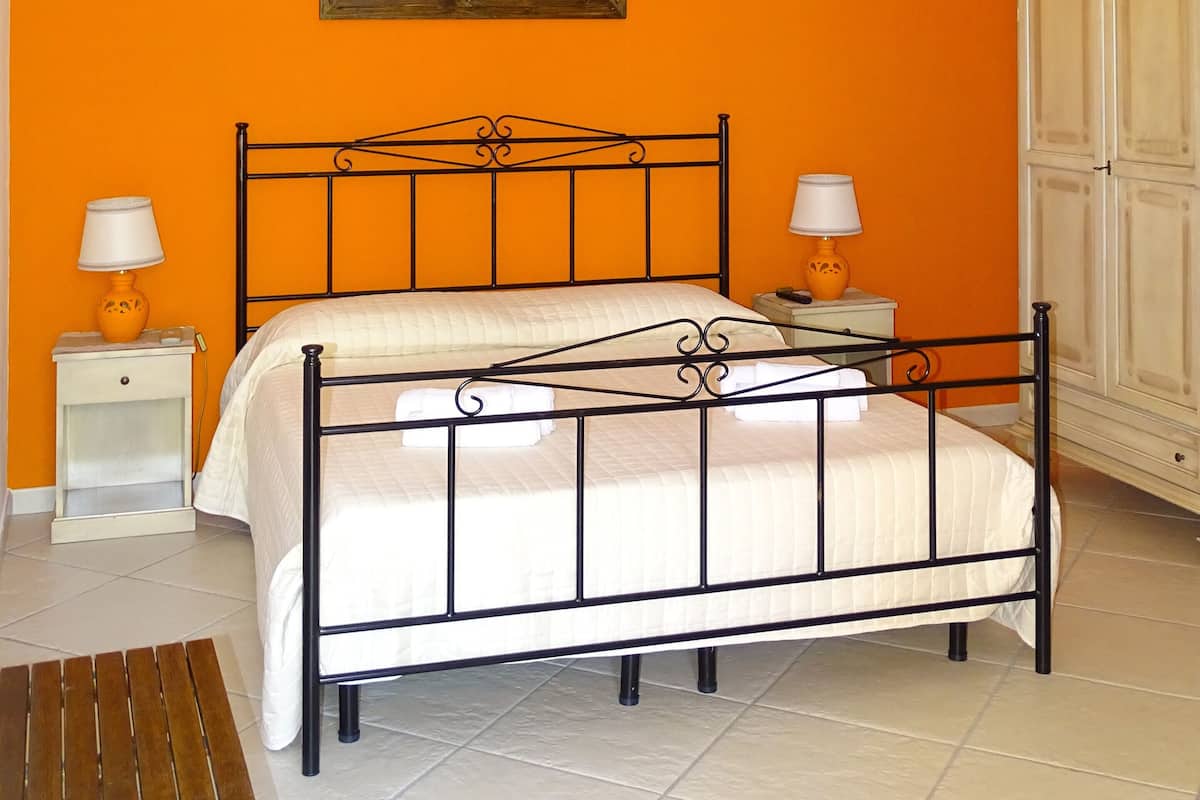 A bedroom with an orange wall, a bed with a black metal frame, two bedside tables with lamps, and a wardrobe.