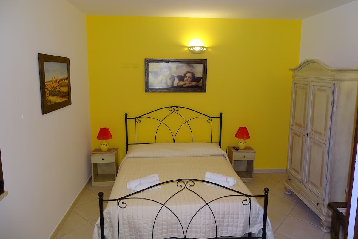A bedroom with a bed, two bedside tables, a nightstand, a wardrobe, a wall-mounted lamp, and two wall-mounted pictures.