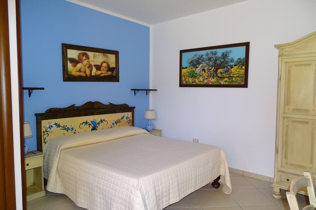 A bedroom with a bed, two wall-mounted lamps, a nightstand, a dresser, and two framed pictures on the walls.