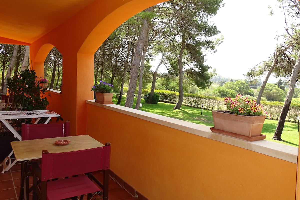 A balcony with a table and chairs, potted plants, and a view of a green landscape with trees.