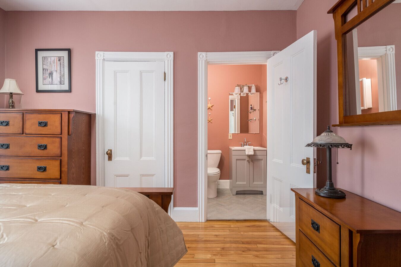 Een slaapkamer met een bed, een kaptafel en een deur naar een badkamer met een wastafel en spiegel.