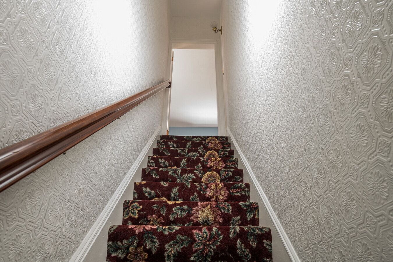 Escada com carpete estampado e corrimão de madeira, levando a uma porta.