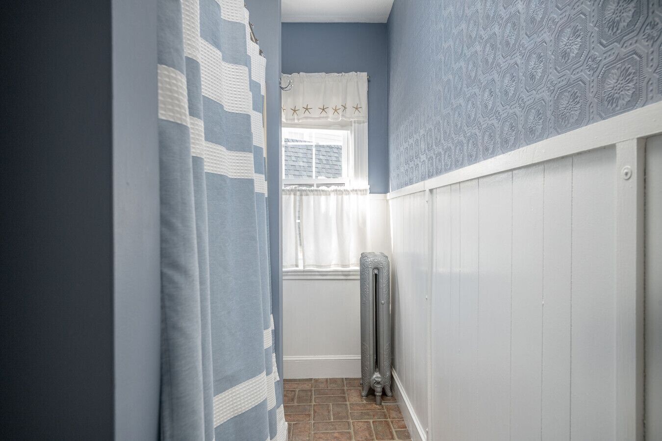 A narrow hallway with a radiator, a window with white curtains, and patterned walls.