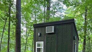 Une petite cabane vert foncé, avec une porte à l’avant, une petite fenêtre et un porche avec des marches menant à l’entrée. Deux plantes en pot sont posées sur le porche.