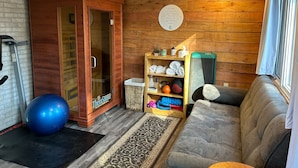 Een knus vertrek met een houten wand, een massagestoel, een kleine fitnessbal, een plank met handdoeken en een waterfles, en een raam met gordijnen.