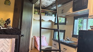 Een compacte keuken met houten kastjes, een zwarte koelkast en een magnetron. Een ladder staat tegen de muur, en een raam zorgt voor natuurlijk licht.