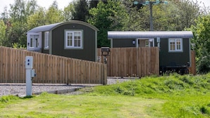 A fenced area with two small, single-story buildings, likely cabins or cottages, with a wooden fence and a utility box in the foreground. - Cabin for 2 With Walks, Wifi, bat and Restaurant (Forfar)