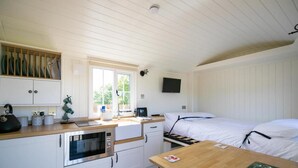 A compact kitchen with a microwave, sink, and wooden counter. There is a bed with white bedding and a small table with a magazine. - Cabin for 2 With Walks, Wifi, bat and Restaurant (Forfar)