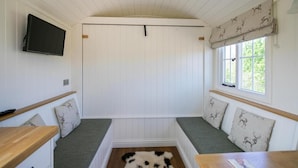 A room with a white paneled ceiling, a large window with a view of greenery, a flat-screen TV mounted on the wall, and a bench with patterned cushions. - Cabin for 2 With Walks, Wifi, bat and Restaurant (Forfar)
