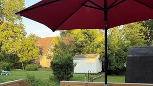 Eine Holzterrasse mit weißen Adirondack-Stühlen, einem kleinen runden Tisch, einem roten Sonnenschirm und einem weißen Schuppen im Hintergrund.