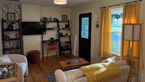 Ein Wohnzimmer mit einer Couch, einem Couchtisch, einem an der Wand befestigten Fernseher, einem Bücherregal und einer Tür mit Fenster.