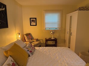 A bedroom with a bed, a chair, a bedside table with a lamp, a window with blinds, and a wardrobe.