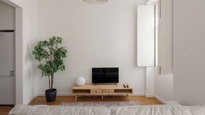 Un salon minimaliste avec un téléviseur à écran plat posé sur une console en bois, une plante en pot et un luminaire blanc au plafond.