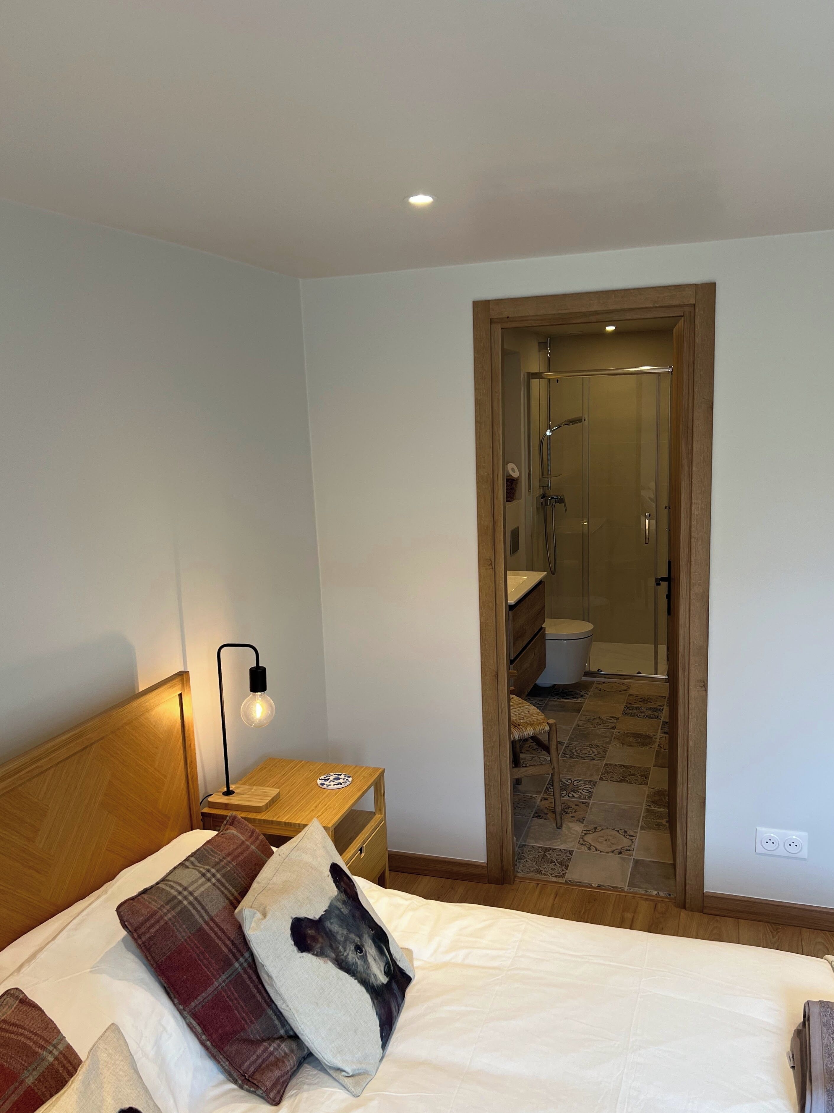 A bedroom with a bed, a nightstand, a lamp, and a bathroom visible through an open door.