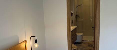 A bedroom with a bed, a nightstand, a lamp, and a bathroom visible through an open door.
