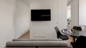 A modern, minimalist room with a flat-screen TV mounted on a white wall, a dining table with chairs, and a grey sofa.