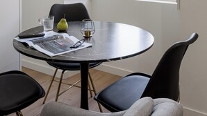 A dining area with a round table, two chairs, a window with open shutters, and a view of greenery outside.