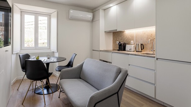 A modern kitchen with white cabinets, a dining area with a gray sofa and black chairs, and a window with a view of greenery.