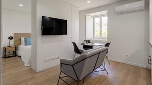 A modern, well-lit room with a bed, a TV mounted on a wall, a dining area with a table and chairs, and a window providing natural light.