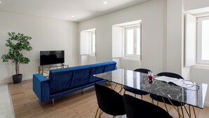 A modern living room with a blue sofa, a flat-screen TV, a glass dining table, and a potted plant.