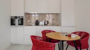 A modern kitchen with white cabinets, a built-in oven, a coffee maker, and a kettle. There is a round wooden table with red chairs and a red apple on it. - The Lisbon Urban Nook Studio (Lisbon)