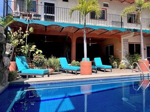 Zona de piscina con sillas de descanso, plantas en macetas y un edificio con balcones.