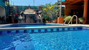 Uma piscina com água azul cristalina, rodeada por plantas em vasos e uma estrutura rochosa decorativa.